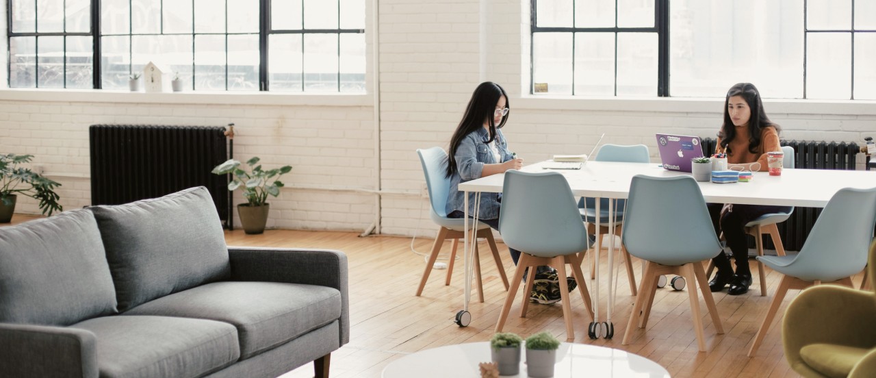 Two working womens in modern office with grey sofa blue chairs and white walls